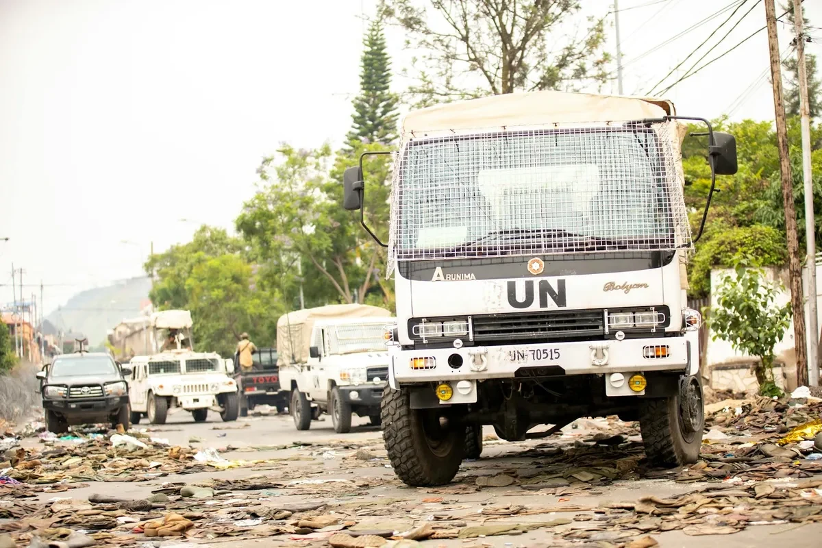 Камиони с хуманитарна помощ и работници разтоварват провизии в лагер в Судан, на заден план се вижда флаг на ООН.