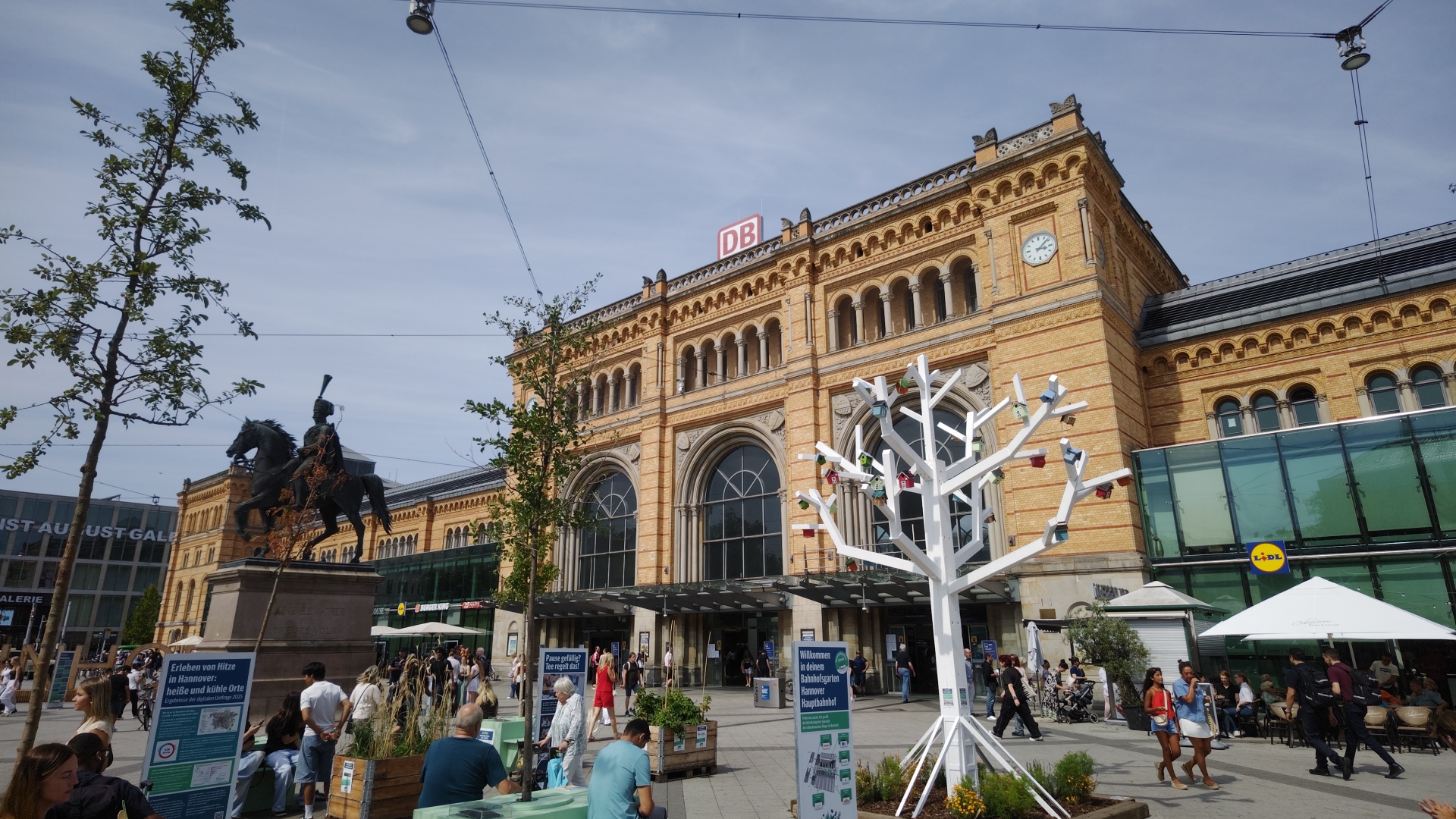 Hannover Station Main entrance with DB logo at top. There is a cast metal statue of a man horseback with a feather in his hat. There are many people sitting and standing around the station.