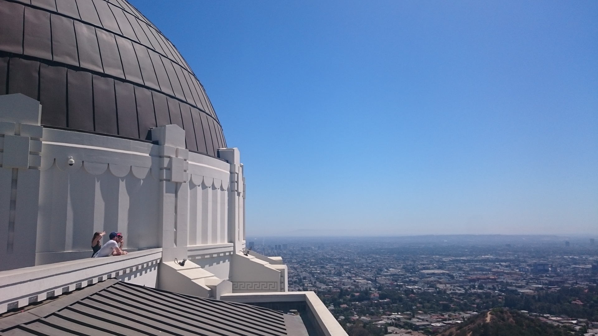 A photo taken from Griffith Observatory in Los Angeles. On the left is the dome and observatory building. The building is a beautiful clean bright white. The some room is made of panels which look like dark bronze. It is a totally clear day with a beautiful clear blue sky and you can see Los Angeles stretch out to the horizon.