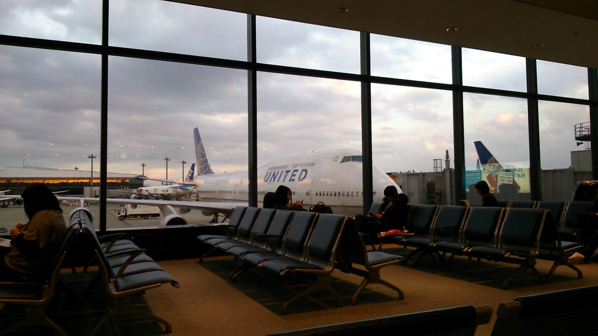 The subject of the photo is a United livery Boeing 747, it is visible behind an airport window and airport seating with travellers waiting.