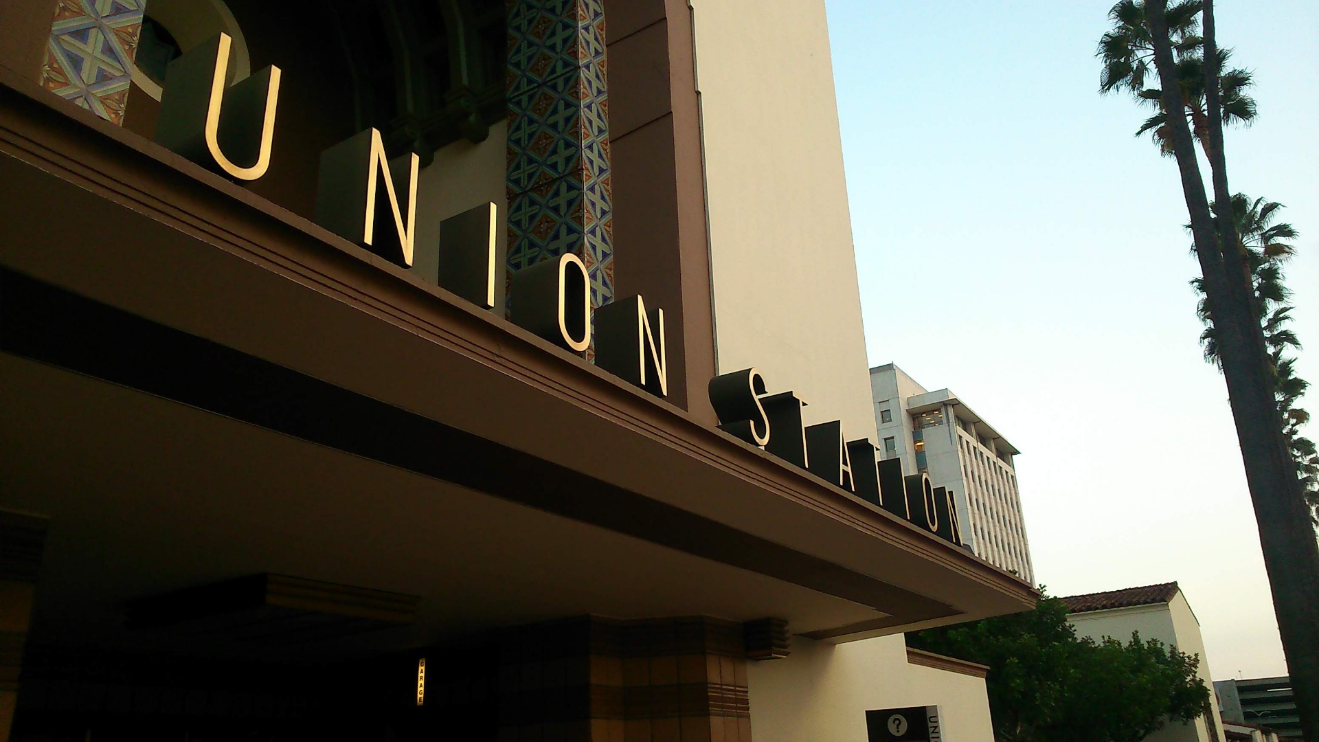 A photo of the art devo tiled grand entrance to union station in Los Angeles. The letters are large San serif block and lit as U N I O N  S T A T I O N. The photo is taken from a perspective shrinking into the horizon. There are palm trees on the right. There are lovely tiles in the station entrance doorway green and orange