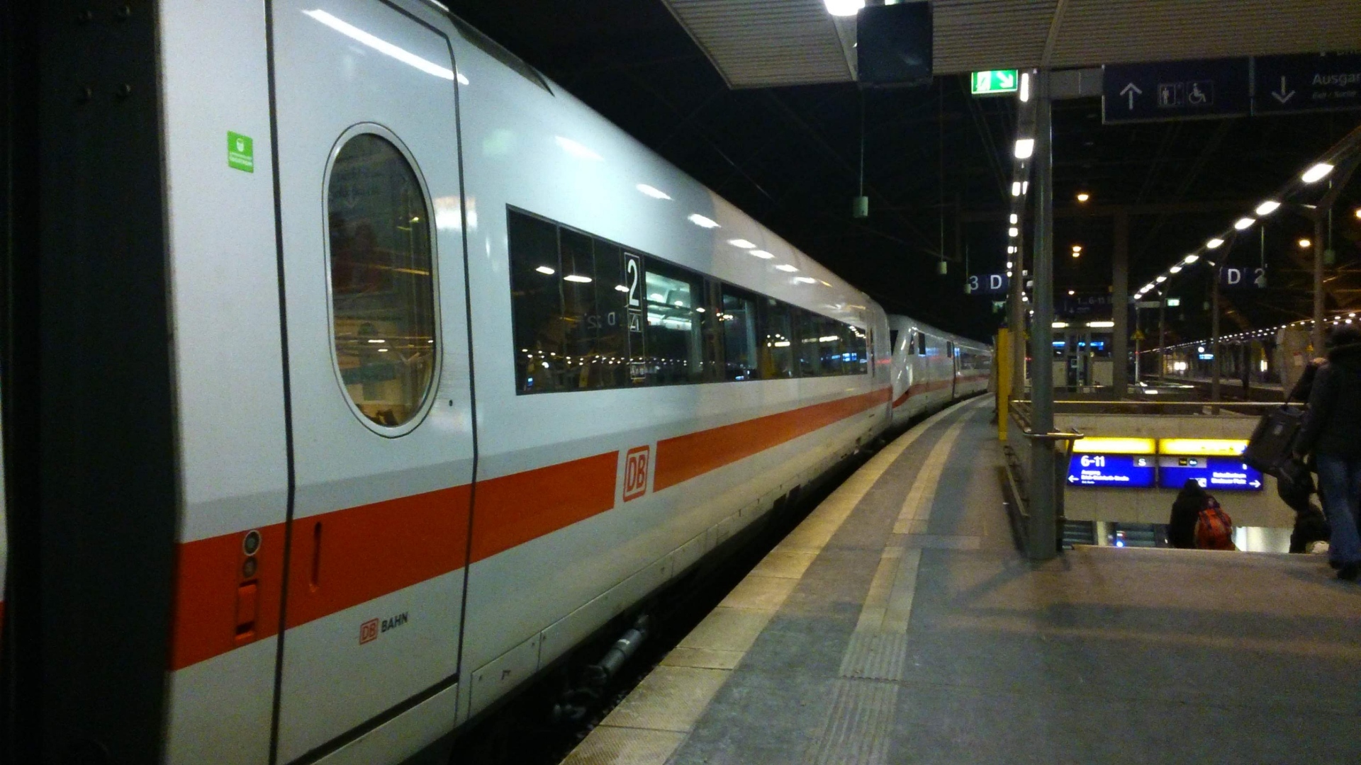 A Deutsche Bahn ICE Train is on a platformin Berlin Station circa 2024, on the right there are stairs down to a passenger concourse with people walking down them. The train door flush with the train surface is visible with a rounded window