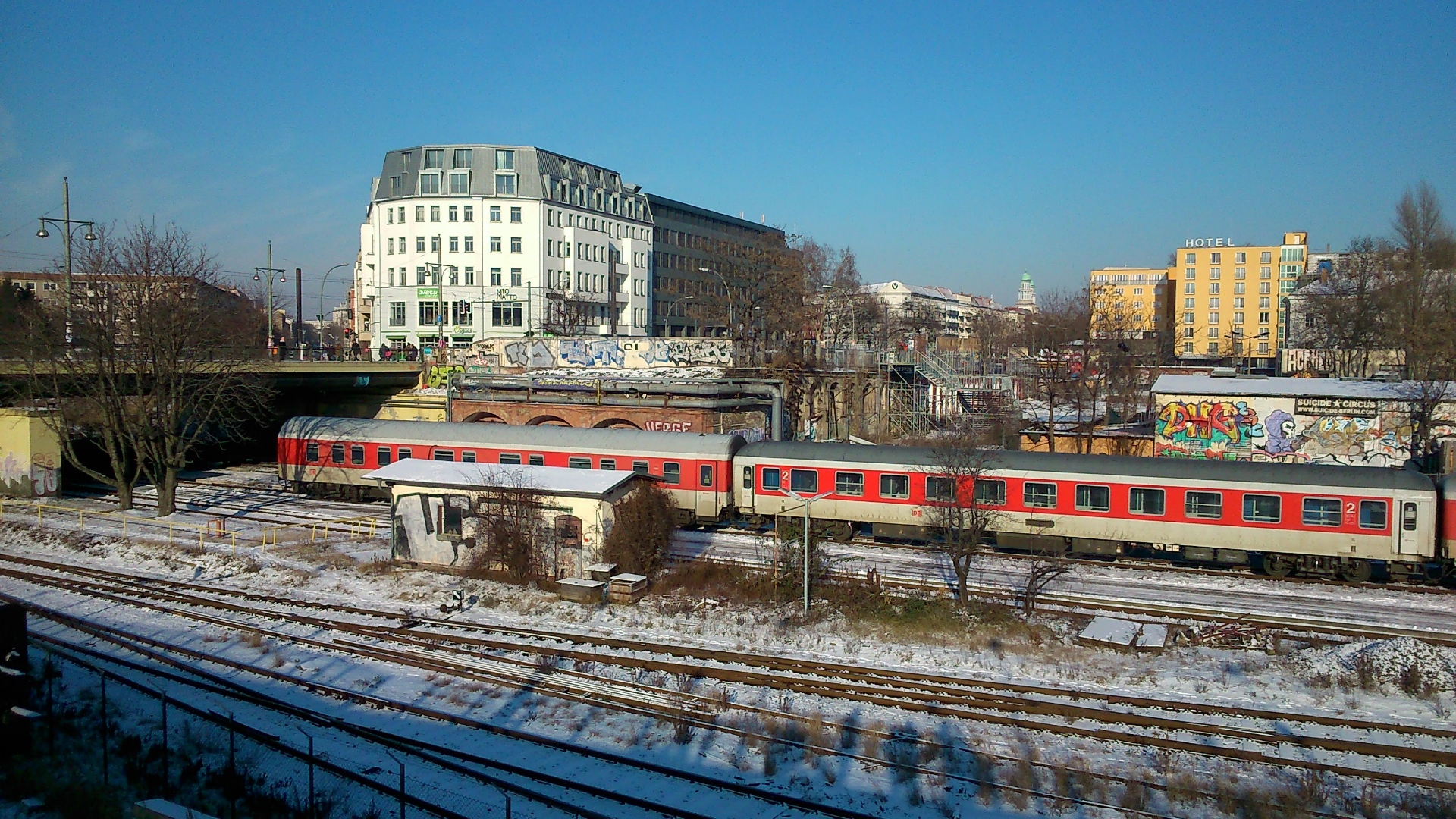A photo of a clear winter day in Berlin. There are red and white Deutsche Bahn train coaches in a rail yard. There is snow on the ground.