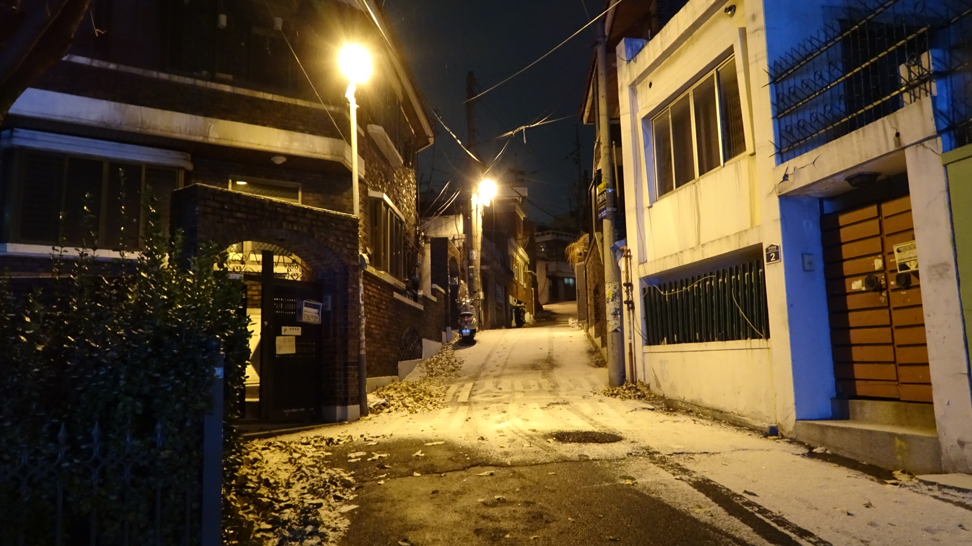 A look up a hill in Seoul. a road goes up the hill and is covered in snow with visible tyre tracks, there is clear pavement in the foreground, there are 2 bright sodium streetlamps illuminating the scene brightly at night all yellow and orange, on the right and left are buildings, the building on the right is clear with a red French style door.