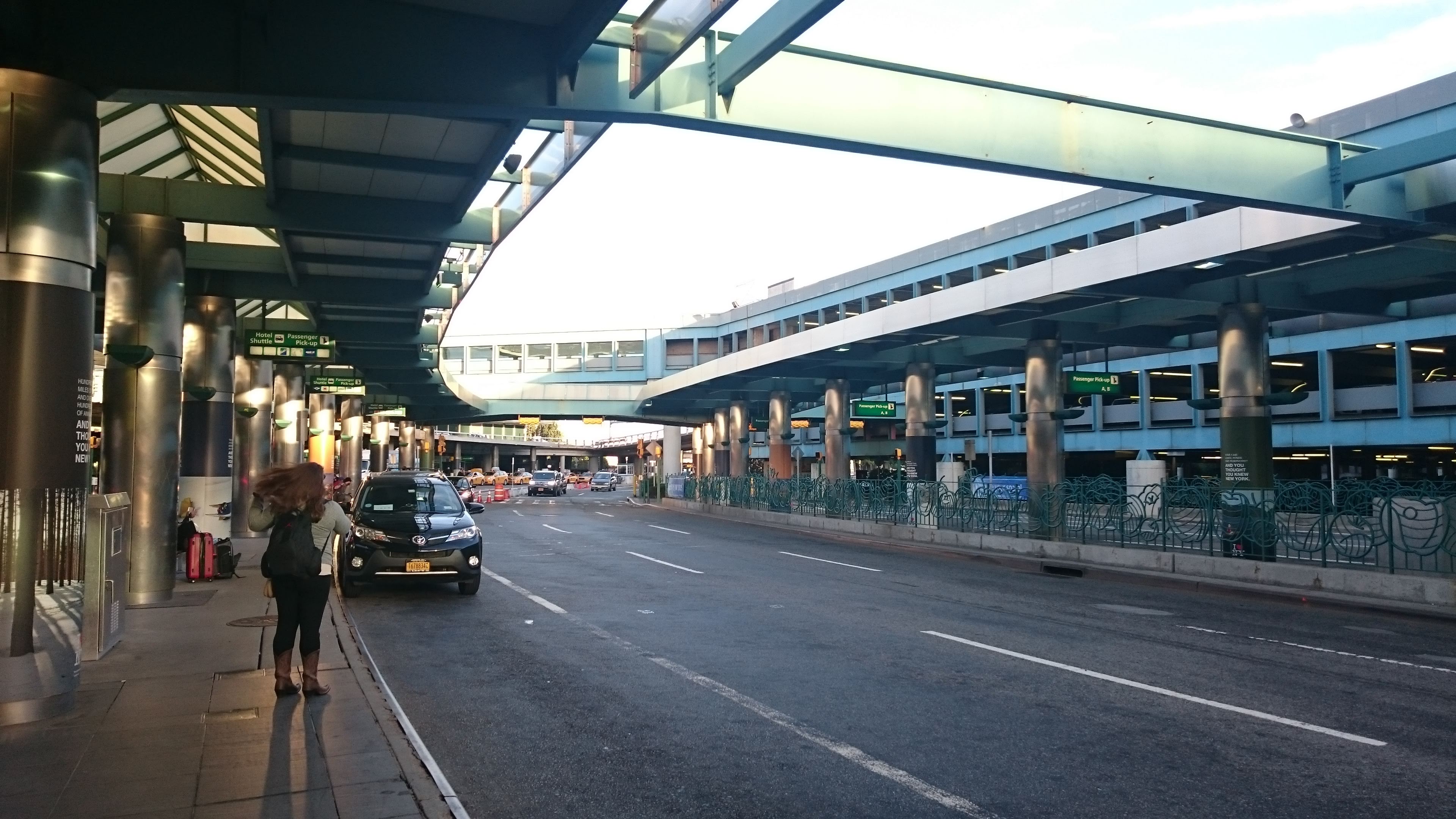 You are standing at sunset at Laguardia terminal B pickup. You are standing on a pedestrian island looking at 3 lanes of traffic. Above you are the terminal overhand roofs and glass beams connecting across to the parking garage. the sun is behind you and shining on the buildings and cars, and shining on the sidewalk. A woman is in motion hailing a taxi to pick her up.