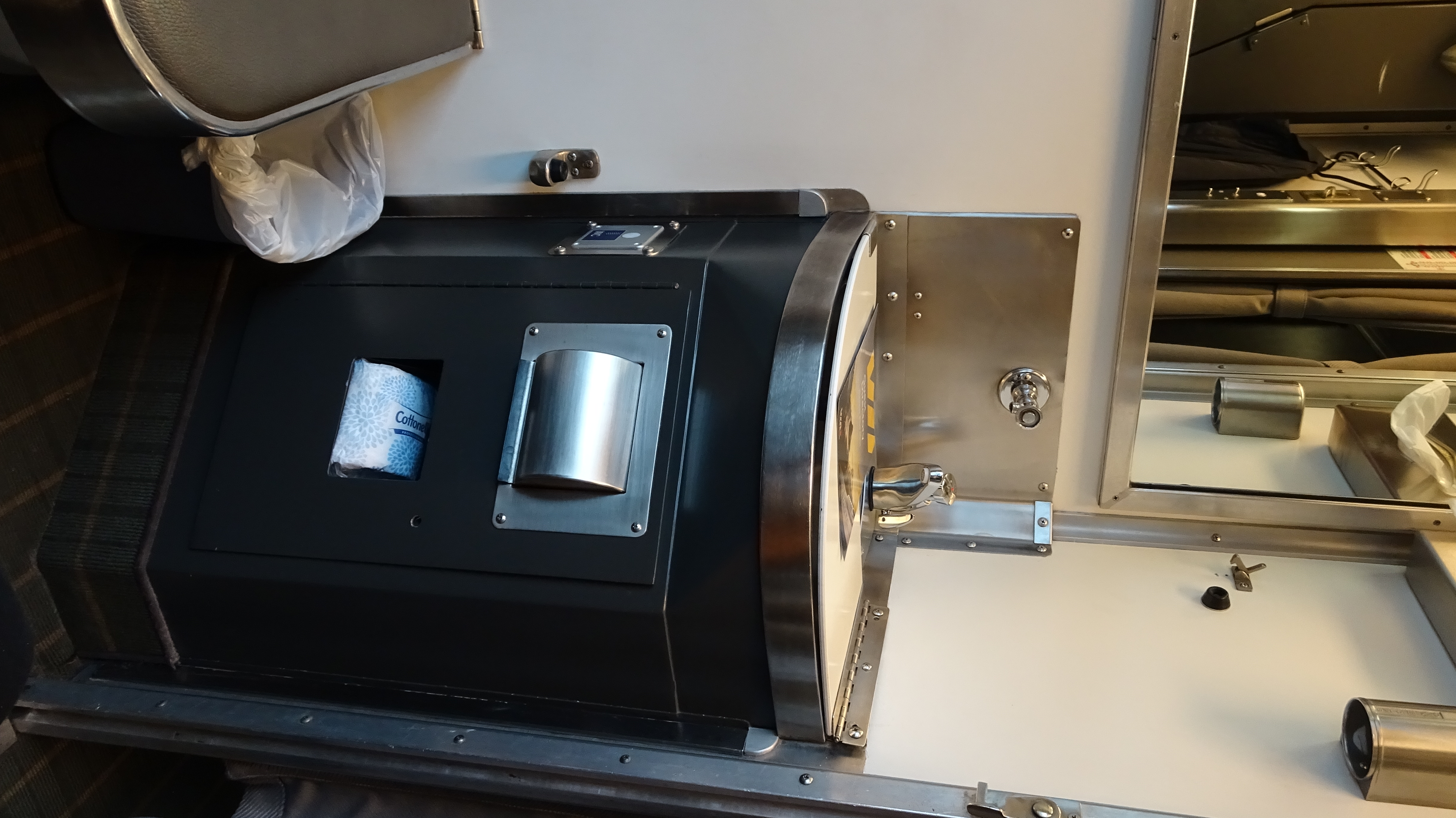 a stainless steel corner sink in a cabin on a train coach, it is supported on top of a black metal podium with a toilet paper dispenser. Above it is a mirror.