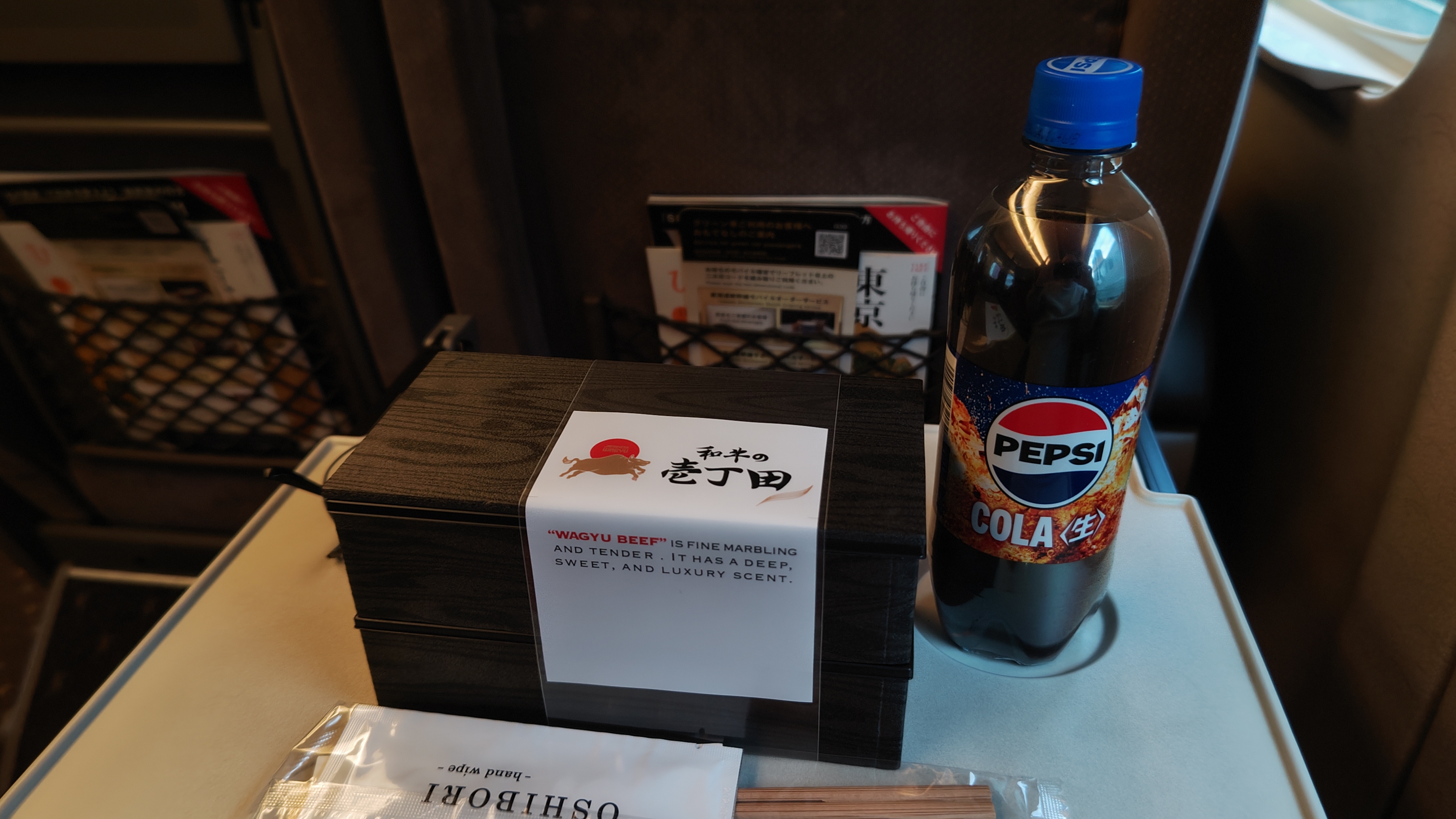 A closed disposable 'eki-ben' bento box containing a wagyu beef meal, with chopsticks infront of it, and pepsi cola to the right
