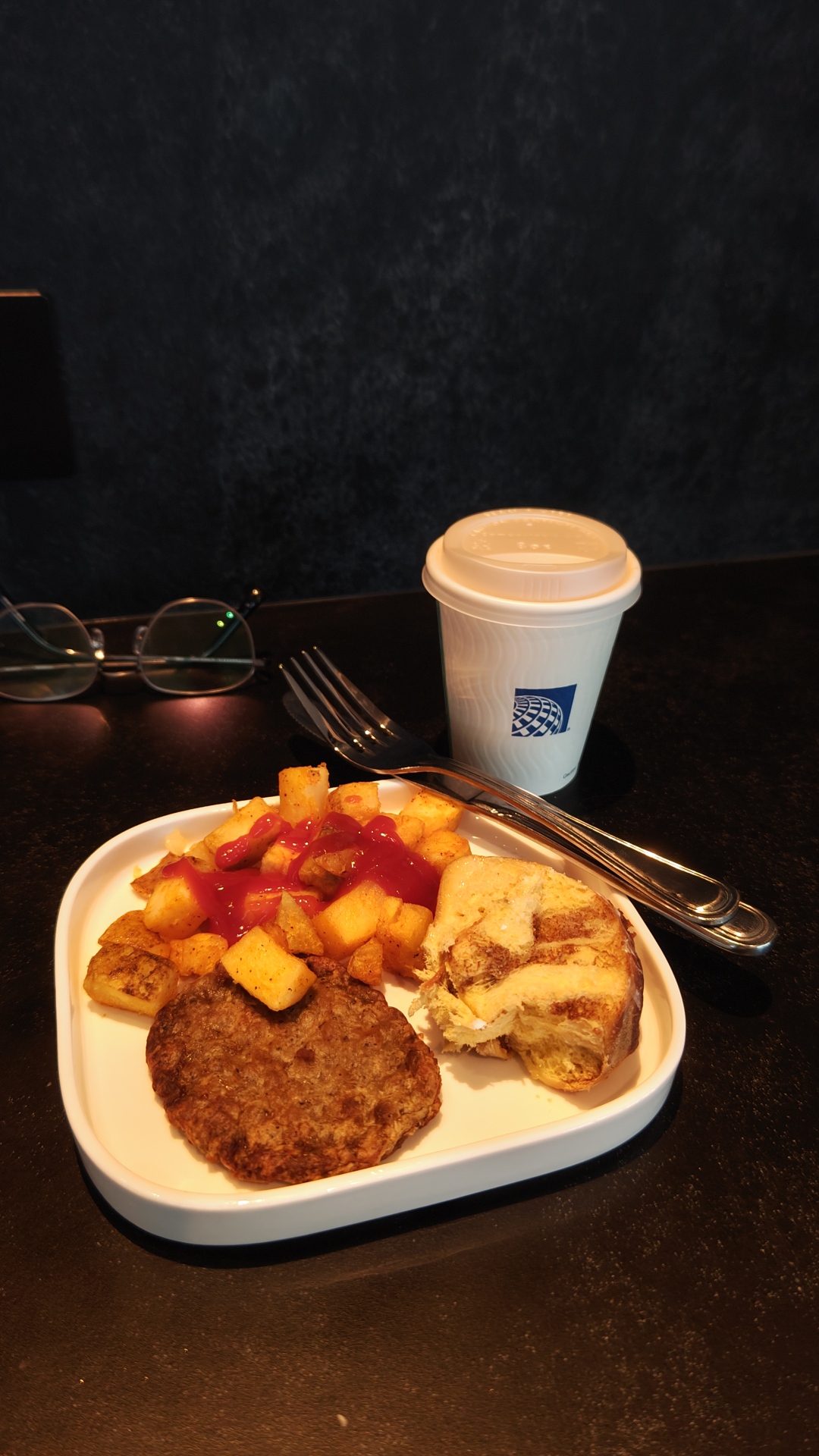 The subject of the photo is a breakfast meal. There are cubed fried potatoes with ketchup on top. There is a slice of breakfast sausage. There is a piece of cinnamon bun roll. It is on a white square plate with rounded corners. There is a fork and knife stacked on the top right corner of the plate. There is a United branded disposable cup of coffee behind the plate, and a pair of glasses.