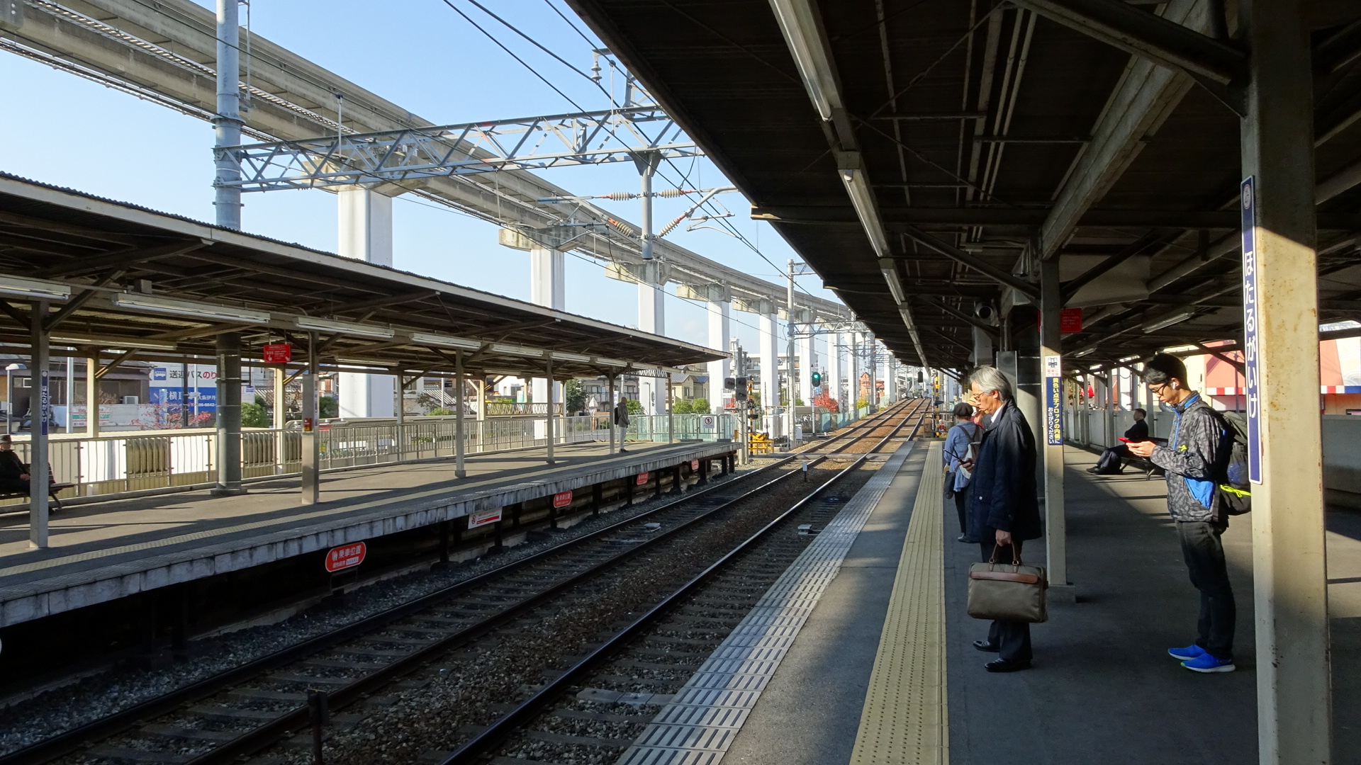 you are standing on the platform in Osaka Japon in a train station and looking down it. There is an older man in his 50s with conservative casual dress and a briefcase looking at his phone absorbed in it. There is a person behind him, a young man perhaps a teenager in casual athletic dress and patterned rain jacket. There are no clouds and it's a clear sky, the sun is behind you shining down the platform.