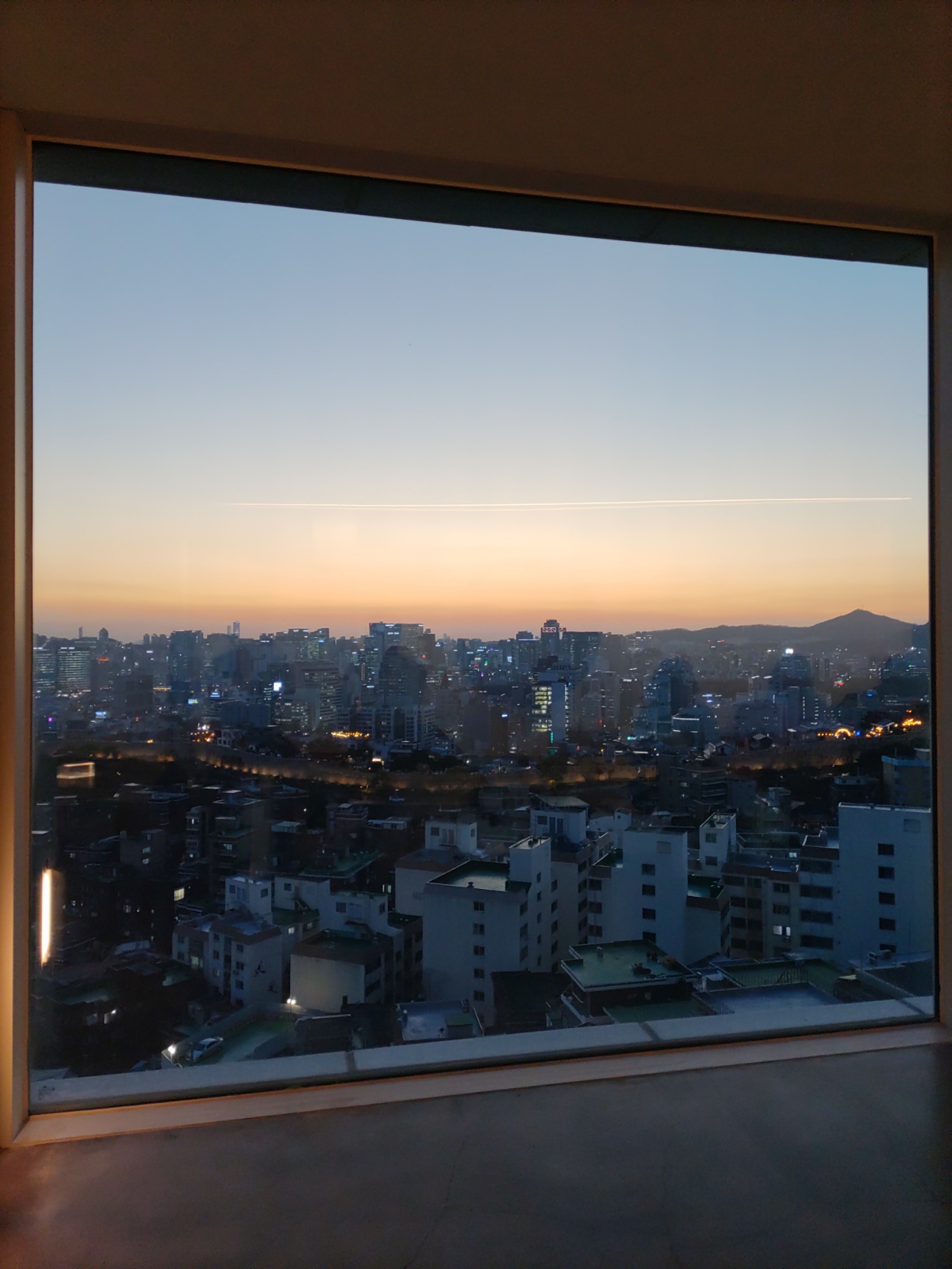 A photo out a large glass picture window before which you can see the dim lights of slow begin to glow in the low dusk as the sun dips below the clouds and mountains at the horizon. The sky is clear besides the line of clouds at the horizon and haze there and a streak in the sky. The ancient city wall is visible and lit with dim yellow light. It separates affordable 20th century apartment housing from the glass modern towers of Seoul. The room is clean concrete with no detail in order to only focus on the incredible view. You are in a cafe on the hillside and looking over the city.