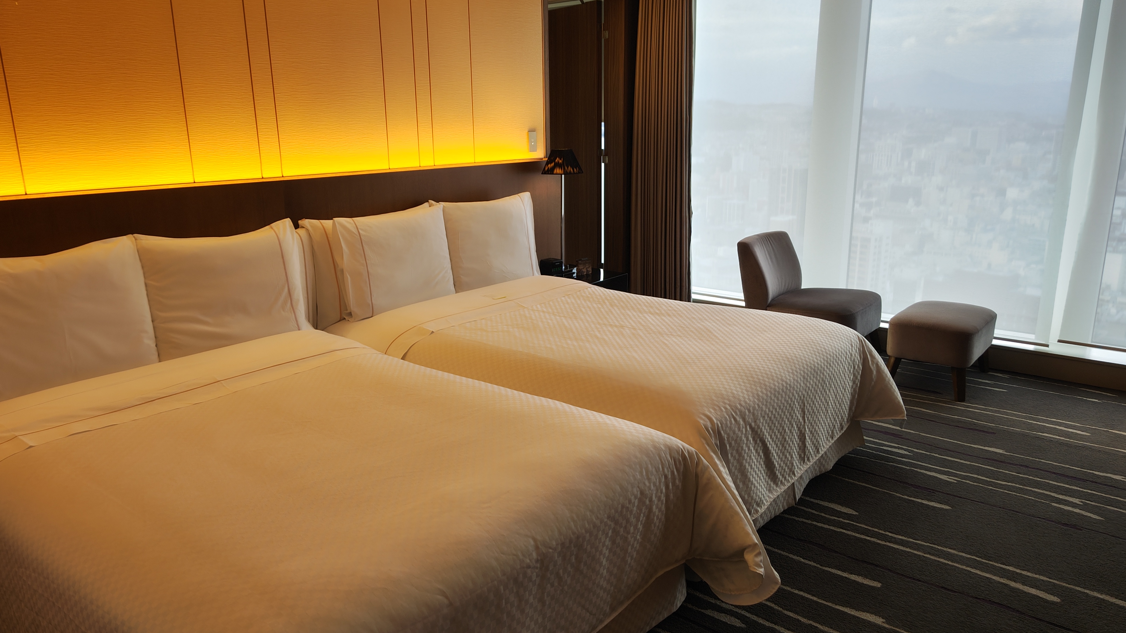 Two double sized beds with white bedding with a soft gentle yellow light from behind, with a chair on the right, in the back are drapes and a window