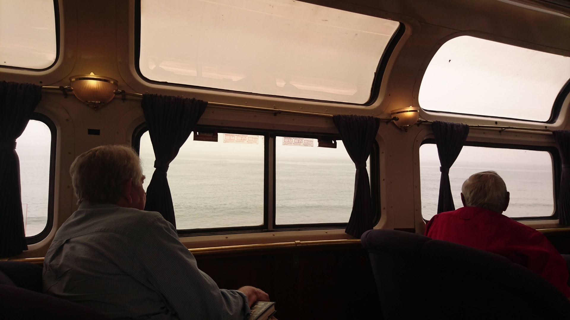 You are sat on an Amtrak superliner observation car. You are facing the wide open curved windows. The cabin is dimly lit by wall sconce lights between the windows. There are two gentlemen sat with their backs to you looking ou the window. Outside the windows it is difficult to make out where the grey sky ends and grey ocean begins, but it is the ocean outside. The lower uncruvwd windows have drapes pulled into bunches on either end.