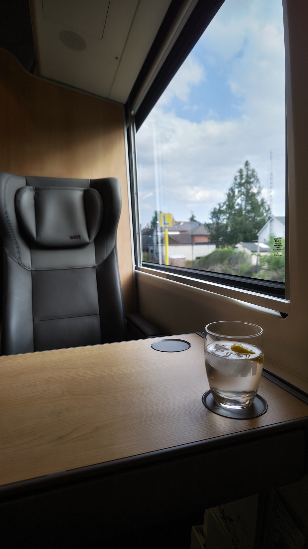 A photo from the semi private seating in the VIA Rail Venture business car. The wooden wall is visible behind the chair across from the photographer. There is a large window on the right and it is daytime with some overcast and blue skies. On the table there is a VIA Rail glass with sparkling water and slice of lemon.