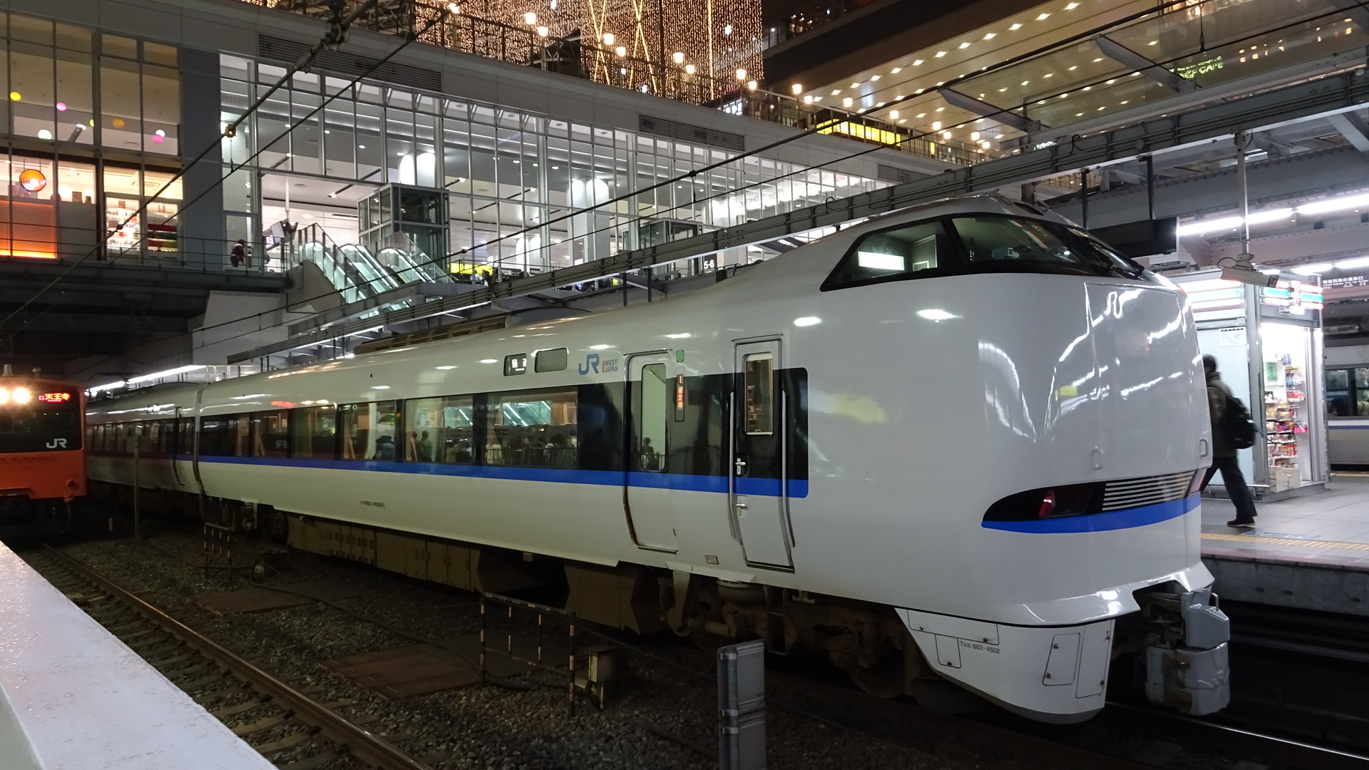 It is a photo of a train in a station. The station is extravagant with many dim lightbulbs providing lighting. There is a glossy white train dominating the photo. It is such a cool looking train. It is perfectly clean glossy white like a brand new Apple device. It has cool IBM blue accents, with black around the windows. The front is totally organically curved with cab windows for drivers on top. It looks futuristic. It is an impressive train in Osaka Japon.