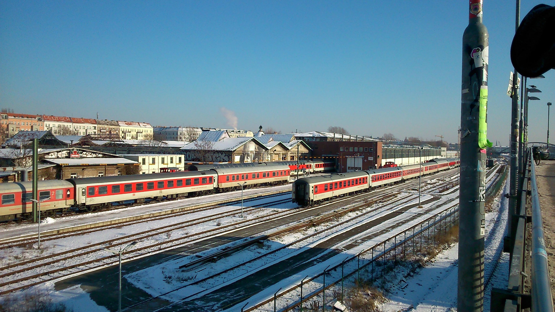 A photo of a clear winter day in Berlin. There are no clouds but a puff of smoke from a chimney. There are red and white Deutsche Bahn train coaches in a rail yard. There is snow on the ground.