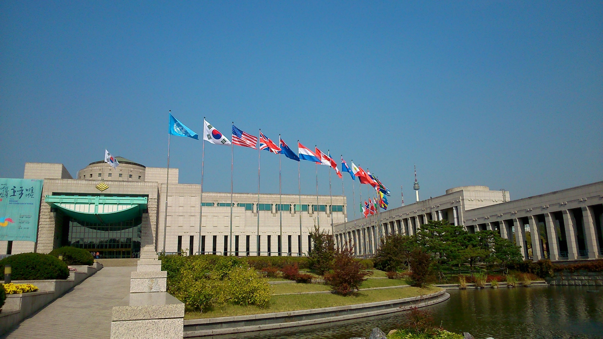 A clear day in Seoul with a perfectly clear sky. There is the impressive war memorial of Korea building. The lawns and gardens and well maintained grass lawns and shrubs. There is a pool of water as a moat. The flags of the nations who fought with United Nations forces in the Korean War / 625 War fly on poles in a line flapping in the wind.