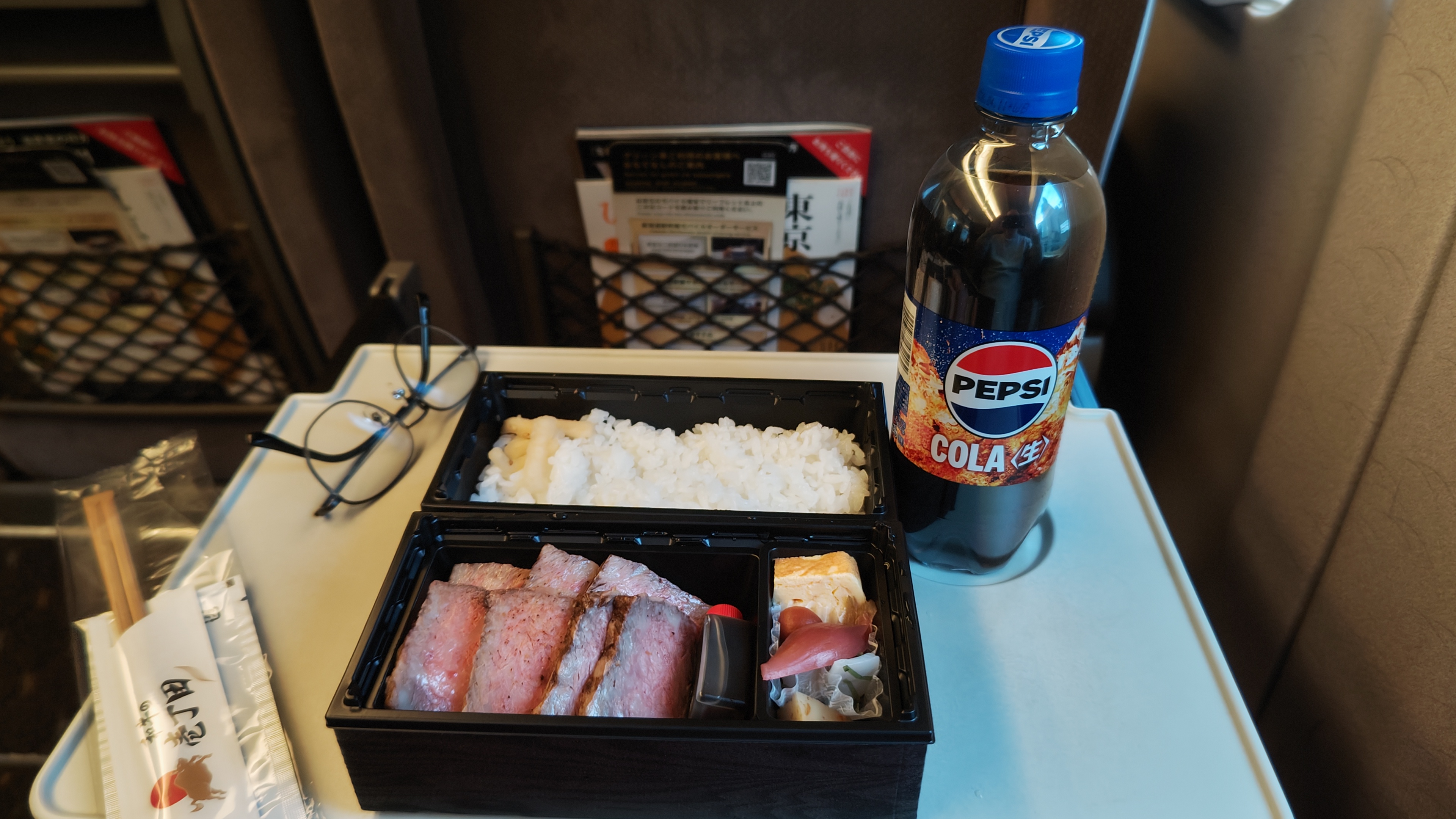 A disposable bento box meal on a shinkansen train, with a bottle of pepsi-cola with a cap. the meal is rice in the top compartment, egg and shallot in the bottom right, and wagyu steak slices in the bottom left.