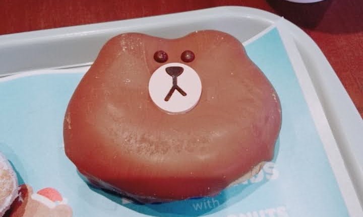 A donut in the shape of the line messenger mascot brown who is a bear with a somewhat despondent and apathetic appearance, it is a chocolate donut with a glossy sheet and decorated with frosting. It sits on a tray with a paper placemat at dunkin donuts Korea