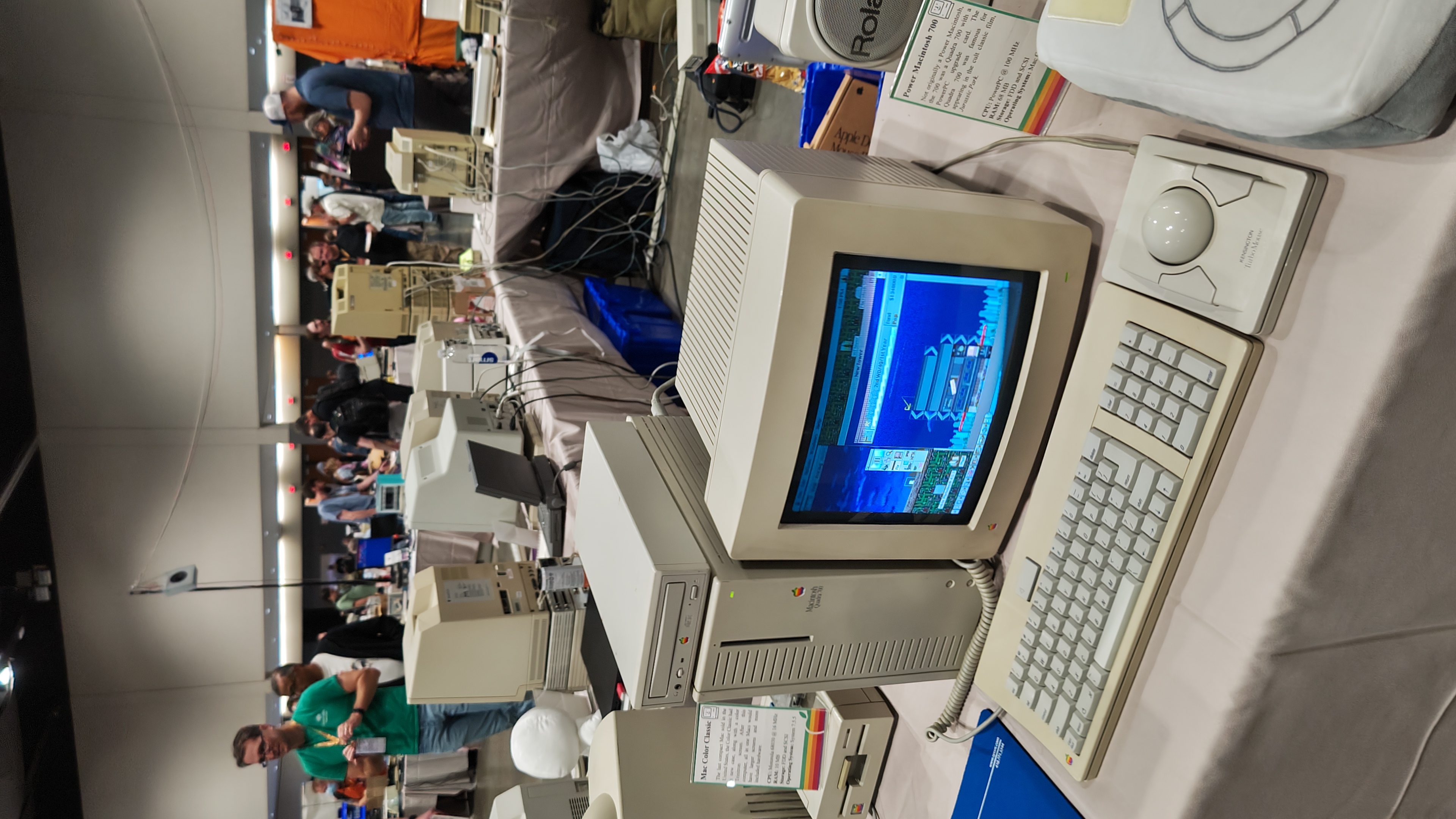 A Macintosh Quadra 700 PC with an Apple CD-ROM drive on top. Next to it is an Apple monitor. It appears to be showing a tower building game. 

A placard to the right with a cute Mac Icon and 4 colour green yellow orange red apple rainbow motif lines at the bottom. It reads:

Title: Power Macintosh 700

Not originally a Power Macintosh, the 700 was a Quadra 700 with a PowerPC upgrade card. The Quadra 700 was famous for appearing in the cult classic film, Jurassic Park. 

CPU: PowerPC @ 100 MHz(sic)
RAM: 68 MB
Storage: FDD and SCSI
Operating System: Mac O(obscured by computer plush)
