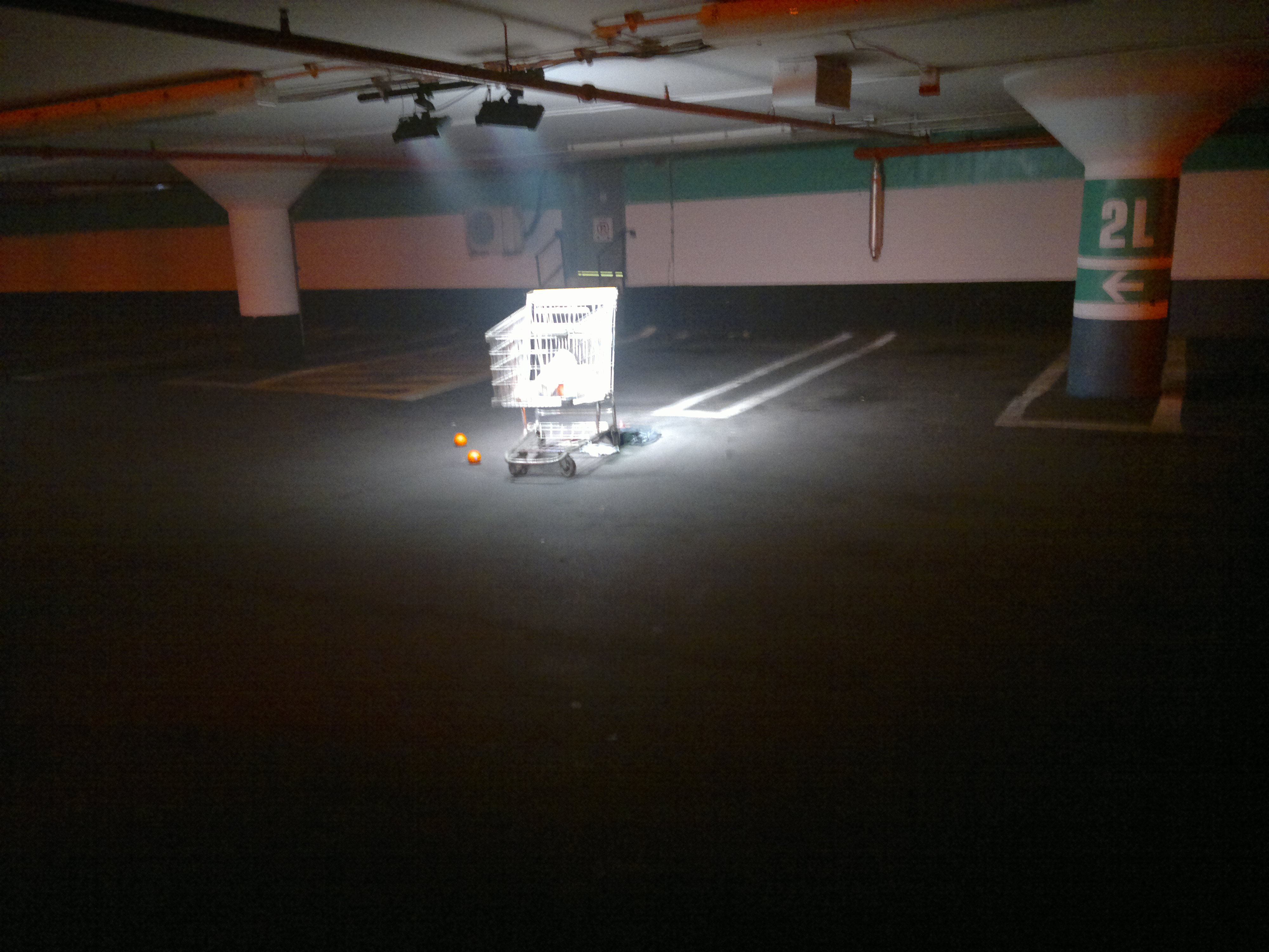 a grocery cart in the parking garage with oranges next to it and it's all washed out and bright with the lamps shining directly on it.