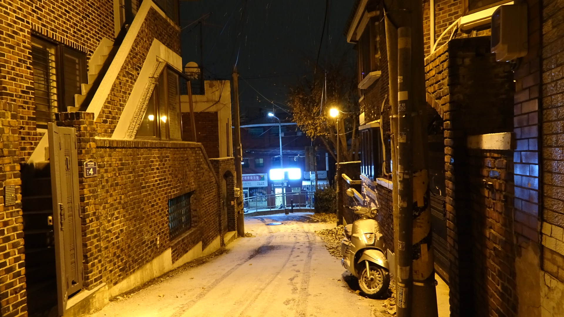 In SeoulA look down a snow covered street with buildings other side at night lit in a orange yellow sodium glow, with car tyre tracks in the snow and bike tyre tracks in the snow partially filled in by freshly fallen snow, and at the foot of the hill a bright white sign